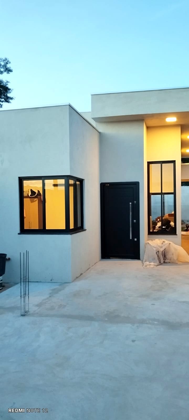 Fachada da casa ao entardecer com porta pivotante em alumínio preto fosco e janela iluminada — projeto real da Linha Suprema executado pela HB Vidros